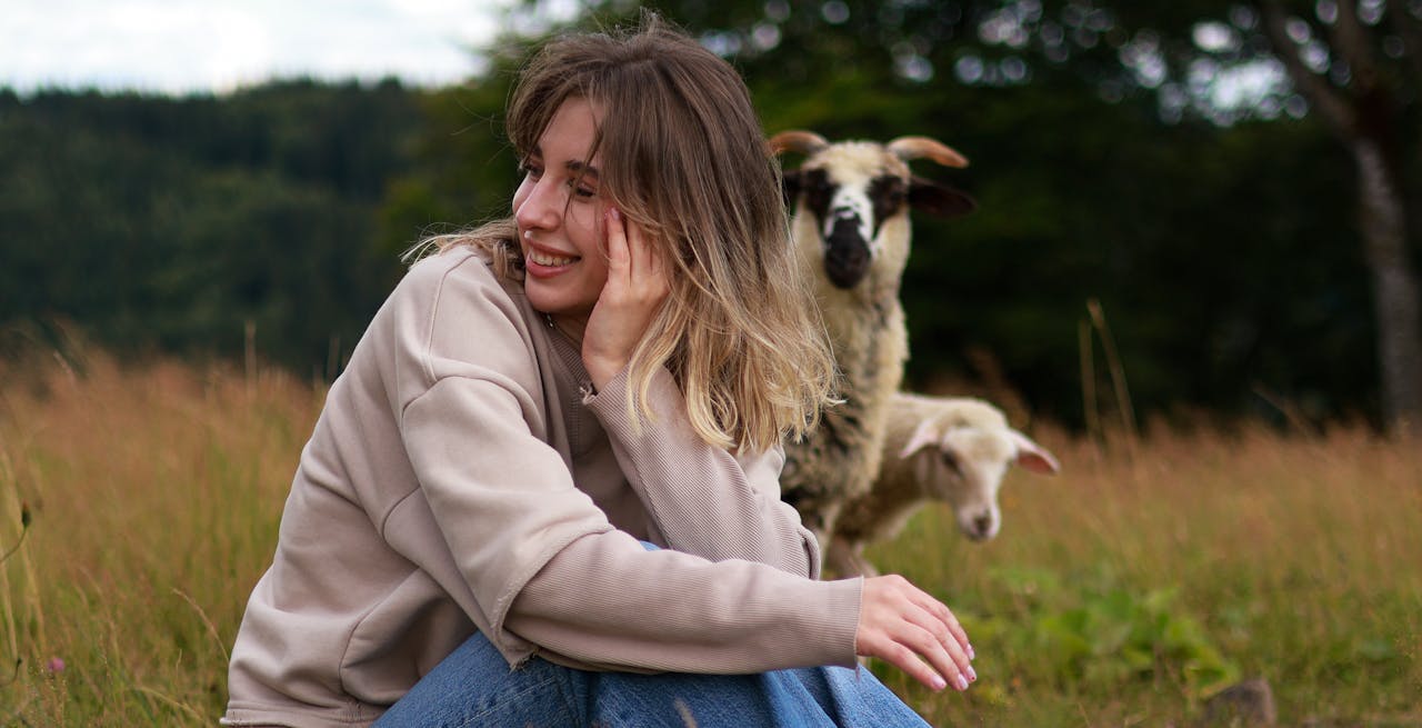 A woman smiles while sitting in a meadow with sheep in the background.